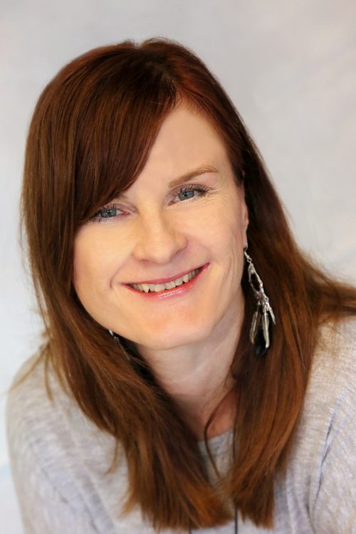 Lächelnde Frau mit langen, rötlich-braunen Haaren, grauem Oberteil und auffälligem Ohrring vor hellem Hintergrund.