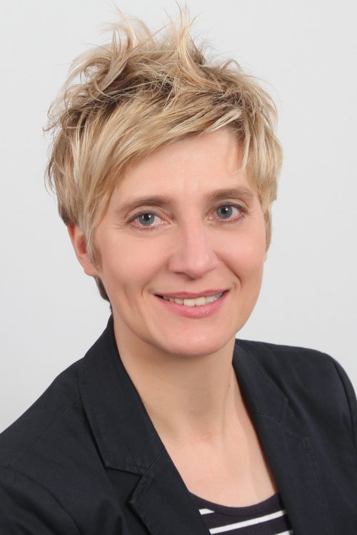 Frau mit kurzem, blondem, fransigem Haarschnitt, dunklem Blazer und gestreiftem Oberteil vor hellem Hintergrund.