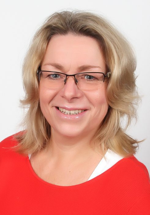 Lächelnde Frau mit schulterlangen, welligen blonden Haaren, Brille und rotem Pullover vor hellem Hintergrund.