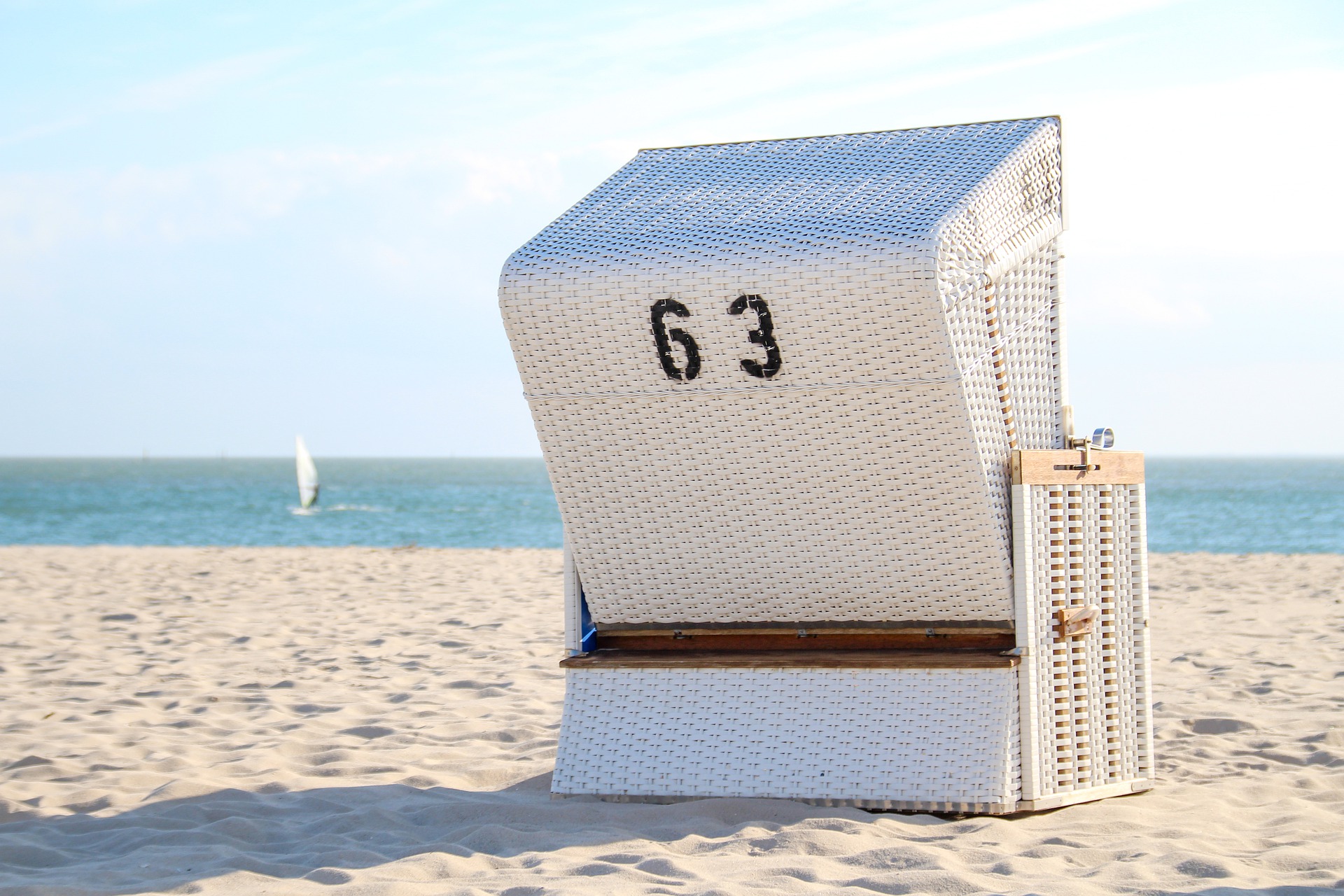 Weißer Strandkorb mit der Nummer 63 steht auf einem sonnigen Sandstrand, im Hintergrund das blaue Meer und ein Segelboot in der Ferne.