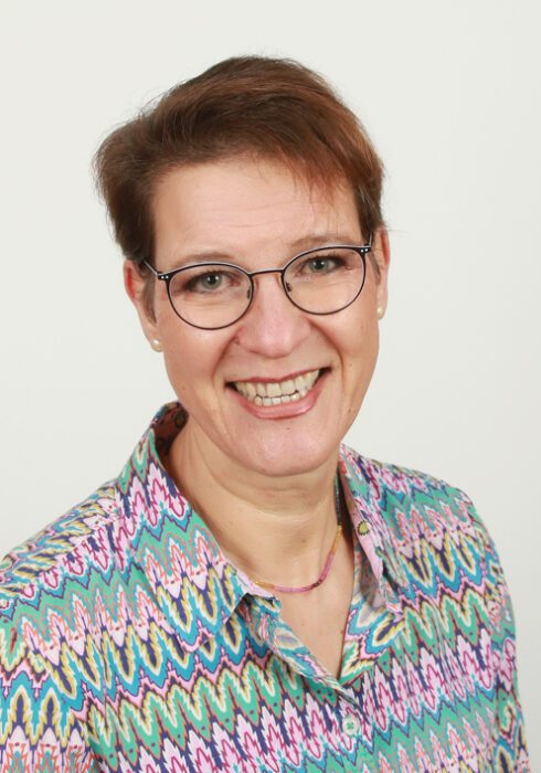 Lächelnde Frau mit kurzem braunem Haar, Brille und bunt gemustertem Hemd vor hellem Hintergrund.