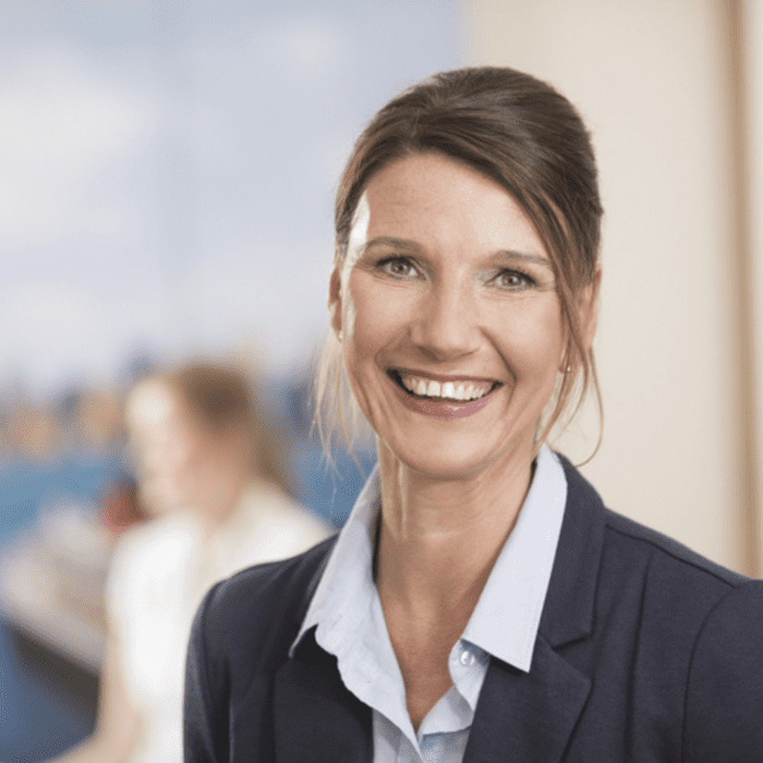 Lächelnde Frau mit dunkelblondem Dutt, hellblauer Bluse und dunkelblauem Blazer in einem modernen Büro mit unscharfem Hintergrund.