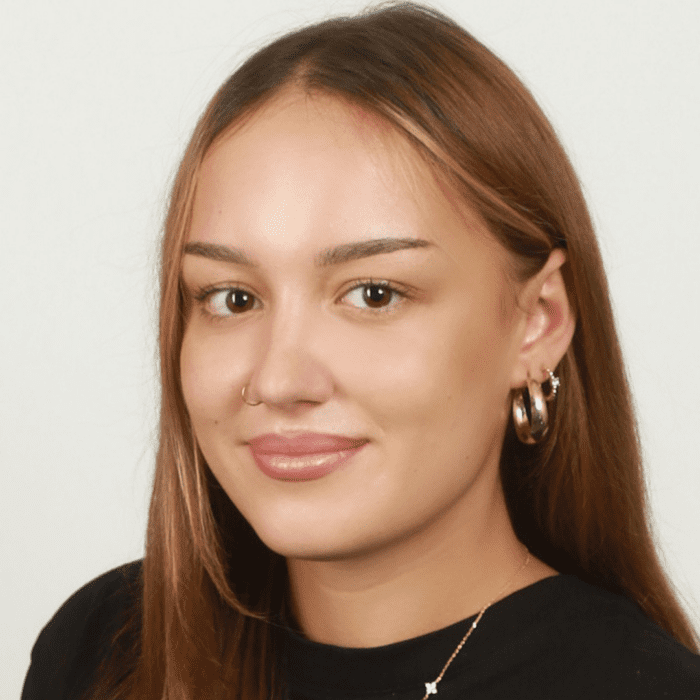 Junge Frau mit langen braunen Haaren, goldenen Creolen und Nasenpiercing vor hellem Hintergrund.