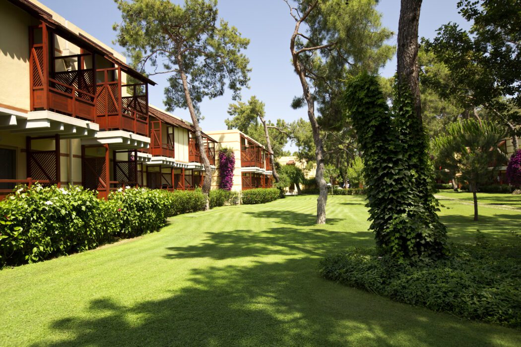 Zweistöckige Ferienhäuser mit roten Holzbalkonen in einer gepflegten Gartenanlage mit grünem Rasen, Bäumen und blühenden Pflanzen unter blauem Himmel.