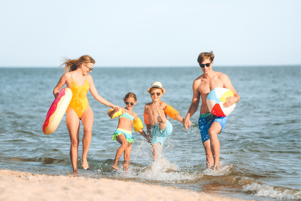 Eine Familie mit zwei Kindern läuft lachend am Strand entlang ins flache Meer, ausgestattet mit Schwimmreifen und Wasserball, bei sonnigem Wetter.