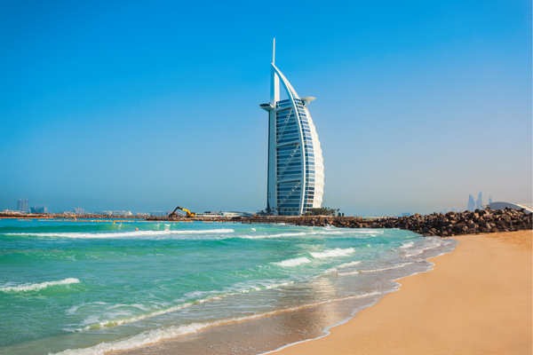 Das Bild zeigt das ikonische Burj Al Arab Hotel in Dubai, das direkt am Strand steht. Der markante, segelförmige Turm erhebt sich stolz gegen den klaren, blauen Himmel. Das türkisfarbene Wasser des Meeres schlägt sanft an den goldenen Sandstrand, während die moderne Architektur des Hotels eine beeindruckende Silhouette bildet. Es vermittelt den Eindruck von Luxus und Exklusivität, in einer der bekanntesten Touristenmetropolen der Welt.