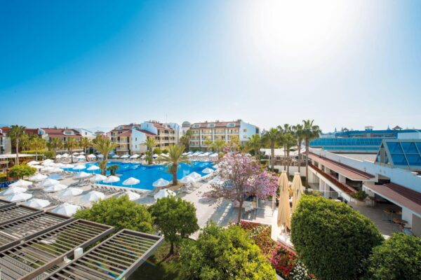 Dieses Bild zeigt einen luxuriösen Hotelbereich mit einem großen Swimmingpool im Vordergrund. Der Pool ist von zahlreichen weißen Sonnenschirmen und Sonnenliegen umgeben, die den Gästen Schatten und Komfort bieten. Im Hintergrund sieht man moderne, mehrstöckige Gebäude mit einem gepflegten, tropischen Garten, der Palmen und bunte Blumen umfasst. Der Himmel ist strahlend blau und das Bild vermittelt eine sonnige, entspannte Atmosphäre, ideal für einen Urlaub. Die Architektur des Hotels und die landschaftliche Gestaltung des Pools unterstreichen das gehobene Ambiente dieses Ortes.