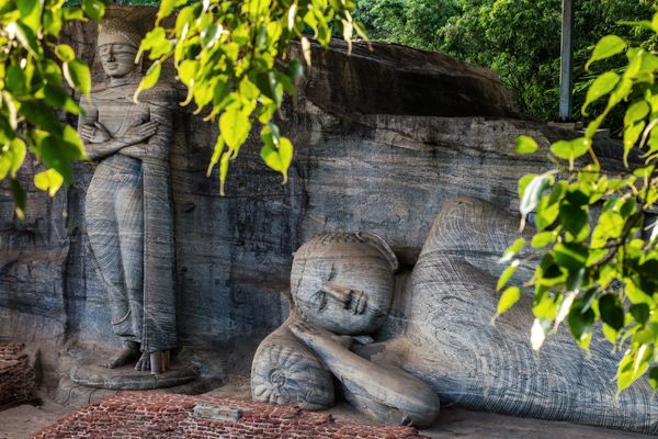 Grosse in Stein gehauene Buddha-Statuen, eine liegend und eine stehend, eingebettet in eine Felswand und umgeben von grünem Laub im Vordergrund.