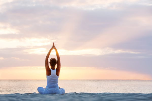 „Person macht Yoga im Lotussitz am Strand bei Sonnenaufgang oder Sonnenuntergang, mit erhobenen Armen in Meditationshaltung, Blick aufs Meer gerichtet.“