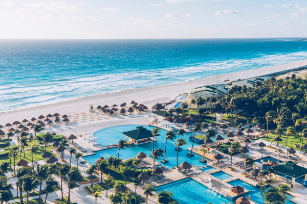 Luftaufnahme eines luxuriösen Strandresorts mit mehreren Pools, Palmen, Sonnenschirmen und Liegen entlang eines weißen Sandstrands mit türkisblauem Meer im Hintergrund.