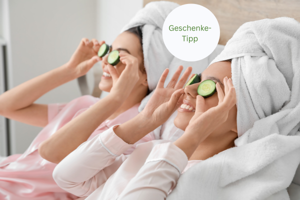 Das Bild zeigt zwei Frauen in einem Wellnessbereich, die entspannen und Gurkenscheiben auf ihre Augen gelegt haben. Beide tragen weiße Handtücher auf dem Kopf und scheinen sich zu amüsieren, während sie eine ruhige Auszeit genießen. Im oberen rechten Bereich des Bildes ist der Text „Geschenke-Tipp“ in einem weißen Kreis zu sehen.