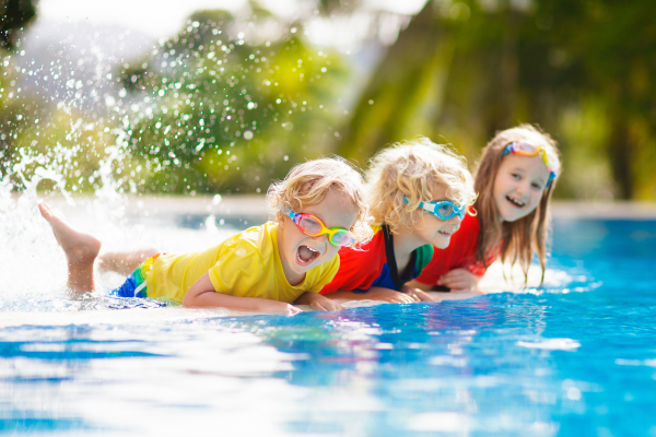 Drei fröhliche Kinder im Pool. Zwei Kinder schwimmen auf dem Bauch, das dritte lacht mit Spritzwasser im Hintergrund. Sie tragen bunte Badebekleidung und Schwimmbrillen.