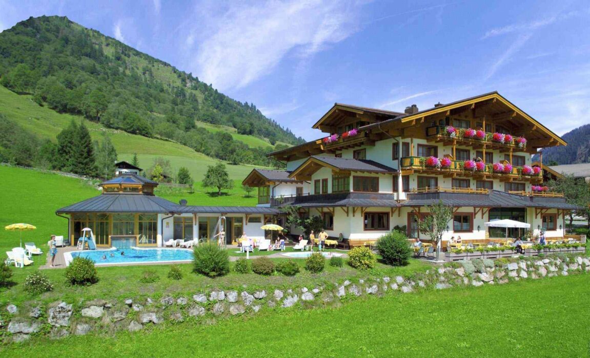 Traditionelles Alpenhotel mit Pool und Sonnenterrasse in grüner Berglandschaft, umgeben von Wiesen, Hügeln und bewaldeten Hängen bei sommerlichem Wetter.