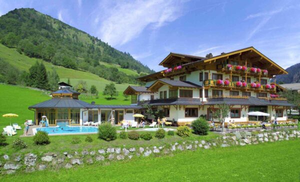Traditionelles Alpenhotel mit Pool und Sonnenterrasse in grüner Berglandschaft, umgeben von Wiesen, Hügeln und bewaldeten Hängen bei sommerlichem Wetter.
