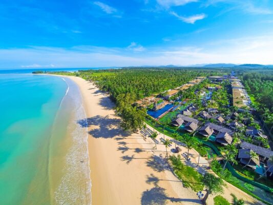 Das Bild zeigt eine atemberaubende Luftaufnahme des Graceland Khaolak Beach Resort in Thailand. Im Vordergrund erstreckt sich der weite, goldene Sandstrand des Resorts, der sanft in das türkisfarbene Wasser des Andamanischen Meeres übergeht. Umgeben von üppigem Grün und Palmen ist das Resort perfekt in die tropische Landschaft integriert. Die weitläufige Anlage bietet eine Vielzahl von luxuriösen Unterkünften und einen schönen Poolbereich, der das paradiesische Ambiente unterstreicht. Im Hintergrund erstreckt sich die Küstenlinie und eine hügelige, grüne Landschaft, die für die Region typisch ist.