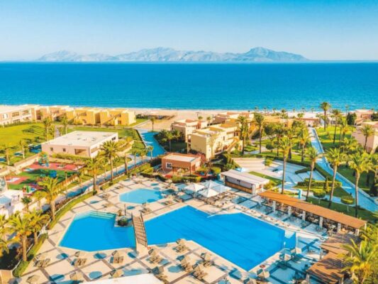 Panoramablick auf ein luxuriöses Strandresort mit mehreren Swimmingpools und palmenbestandenen Gärten, die sich entlang der Küste erstrecken. Im Hintergrund ist das ruhige blaue Meer und eine bergige Insellandschaft zu sehen.