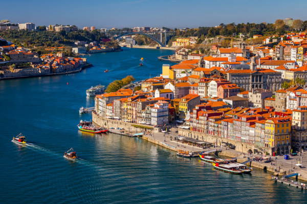 Blick auf die Altstadt von Porto am Ufer des Douro-Flusses mit bunten Häusern, roten Dächern und Booten auf dem Wasser. Im Hintergrund ist die Ponte Dom Luís I zu sehen.