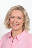 Porträt einer lächelnden Frau mit schulterlangen blonden Haaren, die ein rosafarbenes Hemd trägt und vor einem neutralen Hintergrund steht.