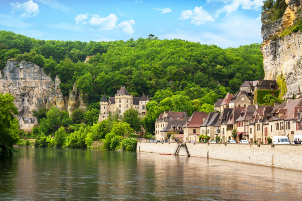 Malerisches Dorf an einem ruhigen Fluss, umgeben von grünen Wäldern und hohen Kalksteinfelsen. Entlang der Uferpromenade stehen historische Häuser mit roten und braunen Dächern, dahinter ein kleines Schloss inmitten der Natur unter blauem Himmel.
