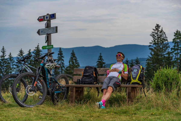 Auf dem Bild ist ein Mann in Fahhradkleidung der auf einer Bank sitzt auf einem berg mit seinem Fahhrad der gerade Pause macht und entspannt.